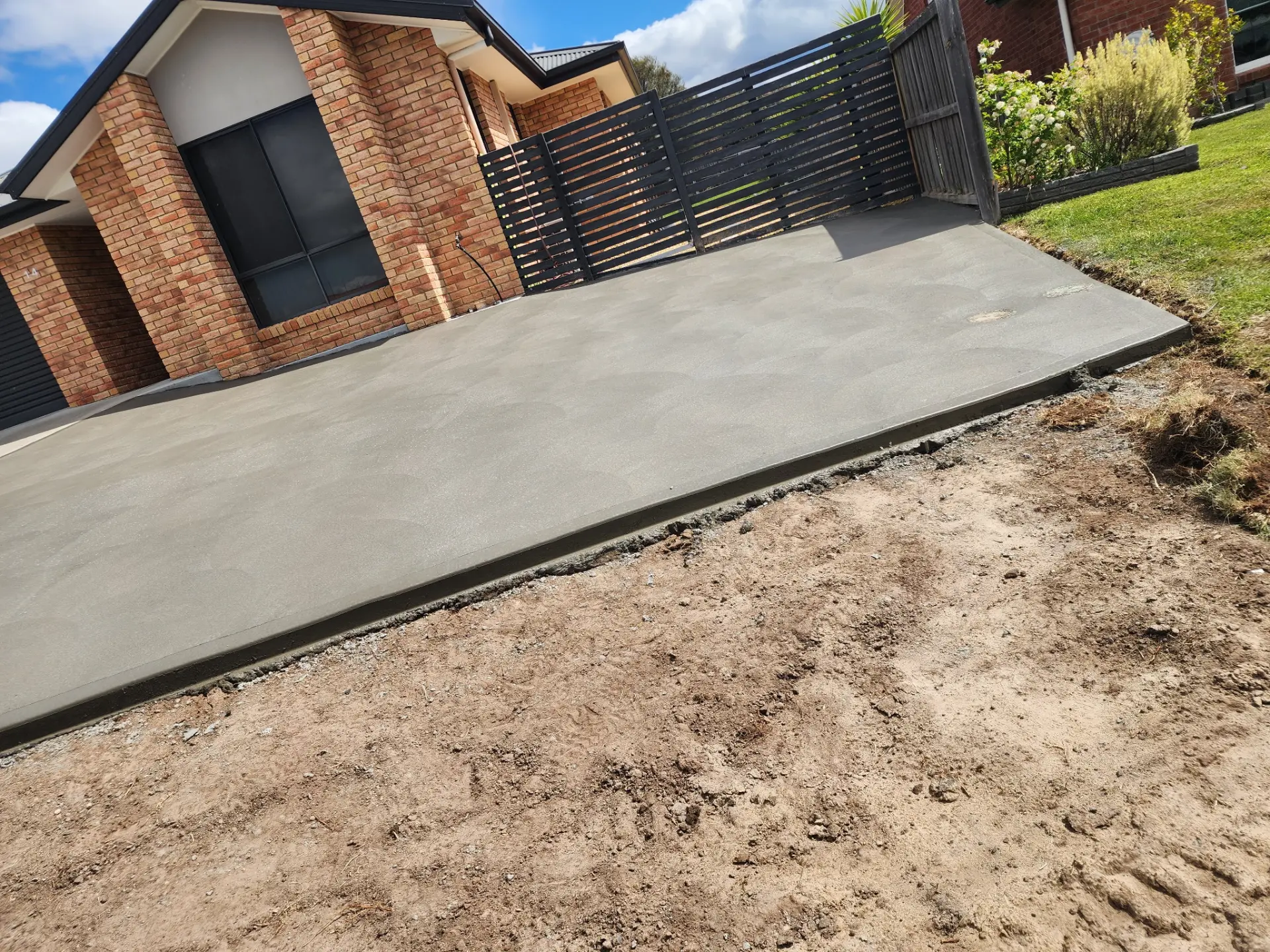 A finished concrete driveway apron beside a brick home with graded earth alongside